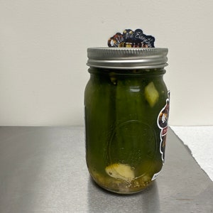 K&ouml;nnte beinhalten: Ein klares Glas mit gr&uuml;nen Gurken und anderen Zutaten, verschlossen mit einem silbernen Deckel. Ein Aufkleber auf dem Deckel lautet "Tickle Me Everything". Ein Aufkleber befindet sich ebenfalls an der Seite des Glases.