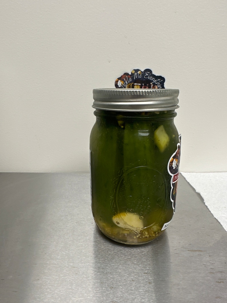 K&ouml;nnte beinhalten: Ein klares Glas mit gr&uuml;nen Gurken und anderen Zutaten, verschlossen mit einem silbernen Deckel. Ein Aufkleber auf dem Deckel lautet "Tickle Me Everything". Ein Aufkleber befindet sich ebenfalls an der Seite des Glases.