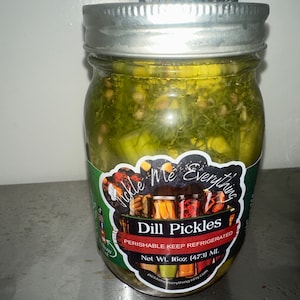May include: A clear glass jar filled with dill pickles, visible herbs, and spices. The jar has a silver lid and a label that reads "Dill Pickles" and "Pickle Me Everything." The net weight is 16oz (473 mL).