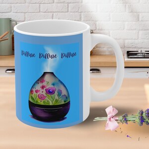 May include: White ceramic mug with a blue background and a colorful illustration of an essential oil diffuser with the text "Diffuse Diffuse Diffuse" printed on the mug.
