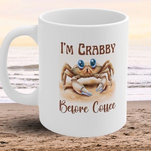 Puede incluir: Taza de café de cerámica blanca con una ilustración de cangrejo marrón y el texto "I'm Crabby Before Coffee".