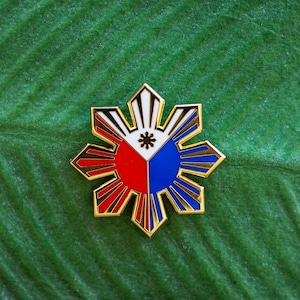 May include: A gold-toned metal lapel pin shaped like an eight-pointed star. The center features a white sun with a small black star, flanked by a red section on the left and a blue section on the right, with white rays extending from the center.