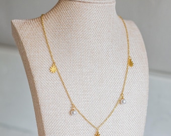 May include: A gold-toned chain necklace featuring alternating small gold flower charms and white spherical beads. The delicate metal chain is arranged in a simple drape, showcasing the evenly spaced floral and pearl-like accents along the length.