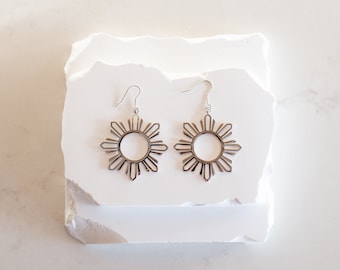 May include: A pair of silver earrings featuring a sun design with eight points. The earrings are hanging from a white surface.