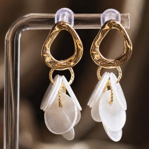May include: A pair of gold-toned earrings featuring an irregular, hammered-texture hoop at the top. Suspended from each hoop is a small, textured gold ring holding multiple white, circular, flat shell-like discs that dangle in a layered cluster.