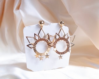 May include: A pair of gold earrings with a circular design and star accents. The earrings are displayed on a white surface.
