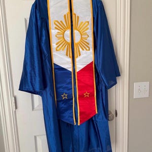 May include: A blue graduation gown with a white and gold sash featuring a sunburst design and the colors of a flag. The sash has a red and blue section with gold stars. The gown is hanging on a white door.