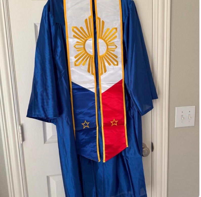 May include: A blue graduation gown with a white and gold sash featuring a sunburst design and the colors of a flag. The sash has a red and blue section with gold stars. The gown is hanging on a white door.