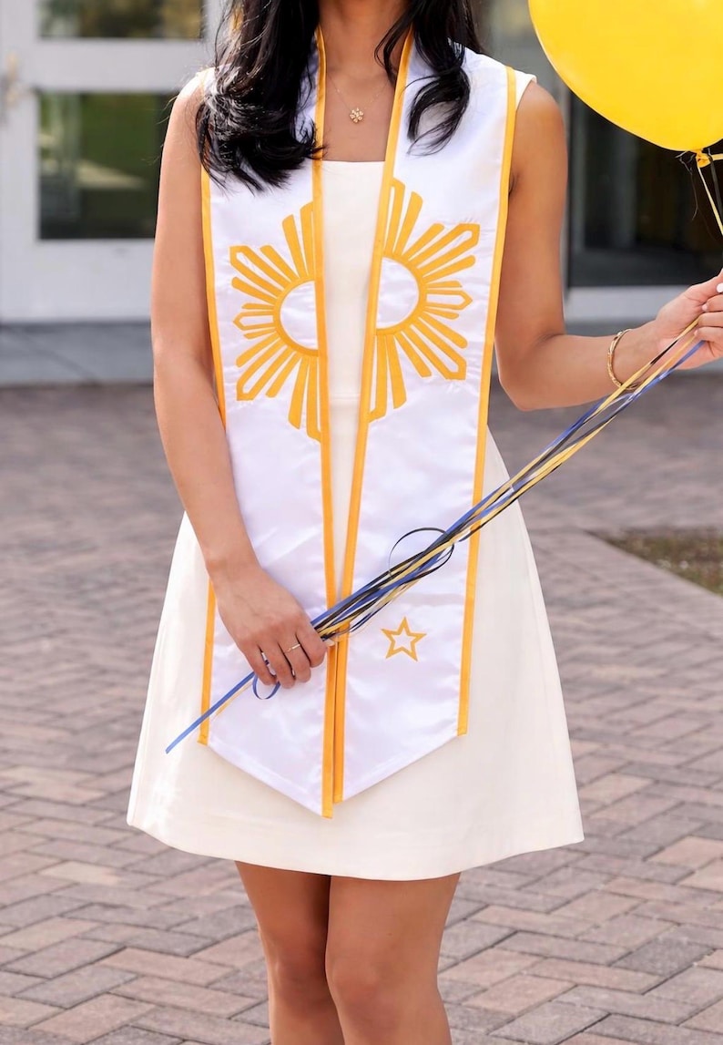 May include: White graduation sash with yellow and gold sunburst design and yellow trim. The sash is worn over a white dress. A yellow balloon and colorful ribbons are held in the hand.