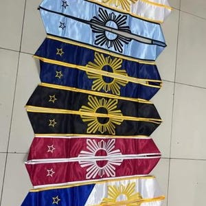 May include: A collection of colorful sashes, each featuring a unique design. The sashes display a sunburst emblem and stars, with variations in color including white, blue, navy, black, red, and a combination of red, white, and blue.