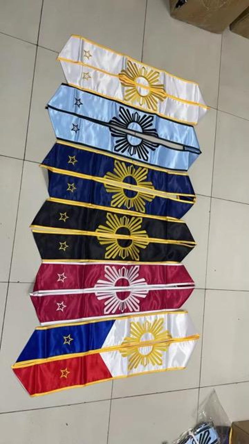 May include: A collection of colorful sashes, each featuring a unique design. The sashes display a sunburst emblem and stars, with variations in color including white, blue, navy, black, red, and a combination of red, white, and blue.