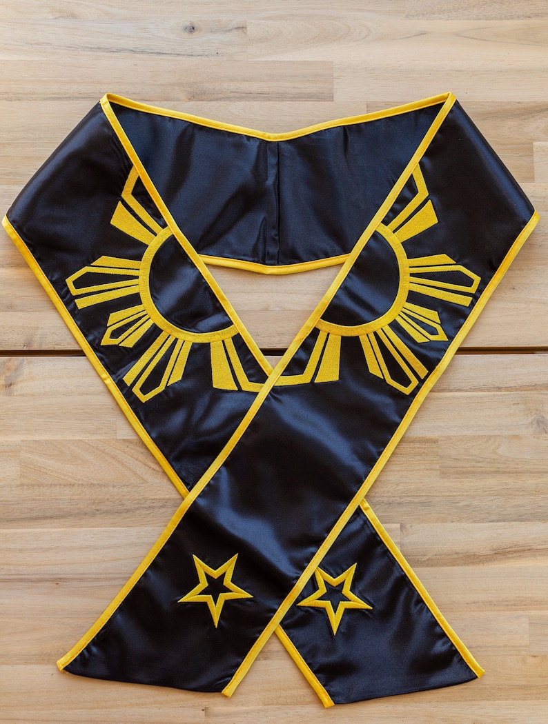 May include: Black graduation sash with yellow trim and embroidered details. The sash features a sunburst design and two stars in yellow. The sash is made of a shiny, satin-like material.