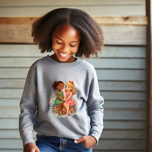 Pode incluir: Um moletom cinza com um gráfico de duas meninas de desenho animado de mãos dadas. Uma menina tem cabelo castanho e usa um vestido verde, a outra tem cabelo loiro e um vestido vermelho. O gráfico está em um fundo floral.