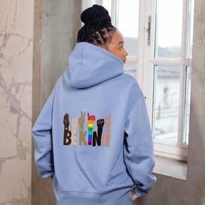 Pode incluir: Uma camisola com capuz azul-clara com um gráfico nas costas. O design apresenta o texto BE KIND, onde cada letra é formada por uma mão a fazer um gesto em língua gestual. As mãos têm vários tons de pele e cores do arco-íris.