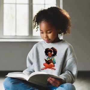 Pode incluir: Uma menina jovem vestindo uma camiseta cinza com um gráfico de uma menina lendo um livro. O gráfico é em estilo de desenho animado e apresenta uma menina com pele marrom, cabelo castanho e uma camisa azul. A menina na foto está segurando um livro e olhando para ele.