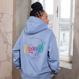 Op de afbeelding: Een lichtblauwe hoodie met een regenboogkleurige afbeelding waarop "You are enough" staat, omringd door woorden als "kind, strong, bold, brave, smart, valued, loved, powerful".