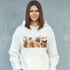 Könnte beinhalten: Ein weißer Hoodie mit einem Aufdruck von vier Herbst-Getränken. Die Getränke sind in verschiedenen Bechern und mit Schlagsahne, Kürbisgewürz und Herbstblättern verziert. Auf einem der Becher steht der Text "Boo Boo".