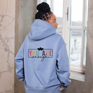Könnte beinhalten: Hellblauer Kapuzenpullover mit dem Aufdruck "YOU ARE enough" auf dem Rücken in einem rechteckigen Design. Der Text ist mehrfarbig, mit zwei schwarzen Herzen über "are". Der Hoodie hat eine Kapuze und einen Kordelzug.