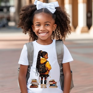 Pode incluir: Uma menina pequena vestindo uma camiseta branca com um gráfico de uma menina carregando livros e uma mochila. O gráfico é de estilo de desenho animado e apresenta uma menina com pele morena e cabelo preto longo. A menina na foto está usando uma mochila cinza e tem um laço branco no cabelo.