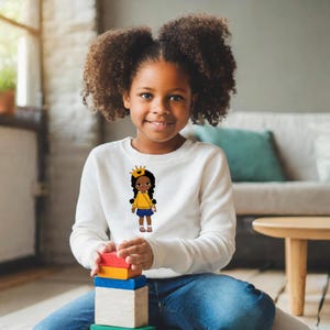 Könnte beinhalten: Ein junges Mädchen trägt ein weißes Sweatshirt mit einer Cartoon-Illustration eines Mädchens, das ein gelbes Shirt, einen blauen Rock und eine goldene Krone trägt. Das Mädchen hält eine kleine gelbe Handtasche. Das Sweatshirt trägt ein Kind, das blaue Jeans trägt.