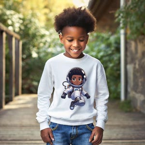 Pode incluir: Camisola branca com um desenho de astronauta de desenho animado. A ilustração mostra uma criança astronauta em um traje espacial e capacete. A camisola é usada por uma criança sorridente usando jeans azuis.