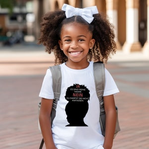 Könnte beinhalten: Ein junges Mädchen trägt ein weißes T-Shirt mit einer schwarzen Silhouette einer Person mit einer Afro-Frisur. Der Text auf dem Shirt lautet "Ja es sind meine Haare Nein du darfst sie nicht anfassen".