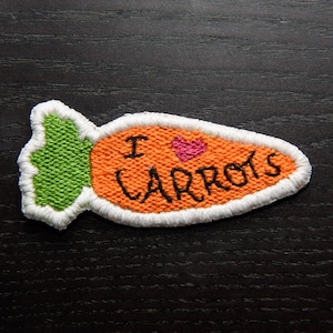 May include: A felt orange carrot with a white outline and green leafy top. The carrot has the text "I love carrots" embroidered on it with a pink heart.
