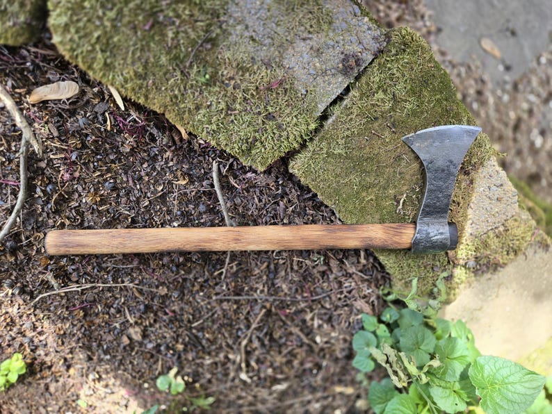 Op de afbeelding: Een handgesmede bijl met een donkere metalen kop en een lange houten steel. De bijl ligt op de grond in de buurt van met mos bedekte stenen en groen. De bijl is een gereedschap om hout te hakken.