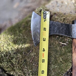 Op de afbeelding: Een metalen bijlkop met een houten handvat rust op een mosachtig oppervlak. Een gele meetlint toont aan dat de bijlkop ongeveer 5 cm breed is. De bijl is een gereedschap om hout te hakken.