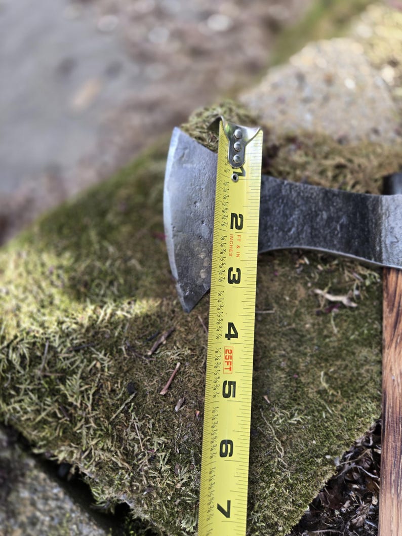 Op de afbeelding: Een metalen bijlkop met een houten handvat rust op een mosachtig oppervlak. Een gele meetlint toont aan dat de bijlkop ongeveer 5 cm breed is. De bijl is een gereedschap om hout te hakken.