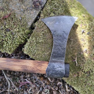 Op de afbeelding: Een handbijl met een houten steel en een stalen blad. De bijlkop is donkergrijs en de steel is lichtbruin. De bijl ligt op een met mos bedekte rots.