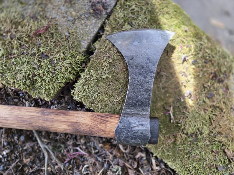 Op de afbeelding: Een handbijl met een houten steel en een stalen blad. De bijlkop is donkergrijs en de steel is lichtbruin. De bijl ligt op een met mos bedekte rots.