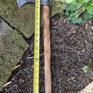 Op de afbeelding: Een handbijl met een donkere metalen kop en een houten steel. De bijl wordt gemeten met een gele rolmaat, met een totale lengte van ongeveer 45 cm.