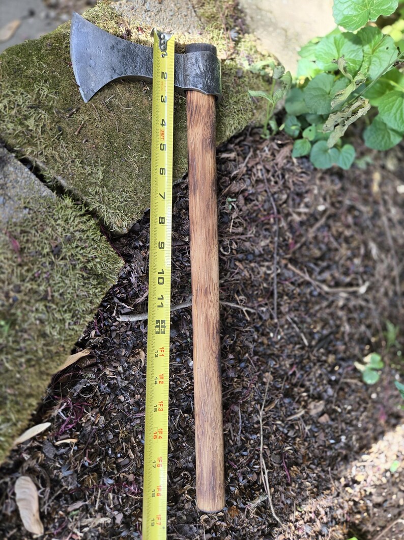 Op de afbeelding: Een handbijl met een donkere metalen kop en een houten steel. De bijl wordt gemeten met een gele rolmaat, met een totale lengte van ongeveer 45 cm.