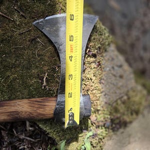 Op de afbeelding: Een handbijl met een donkere metalen kop en een houten steel. De bijlkop is ongeveer 15 cm lang, gemeten met een gele rolmaat. De bijl ligt op een bed van groen mos.