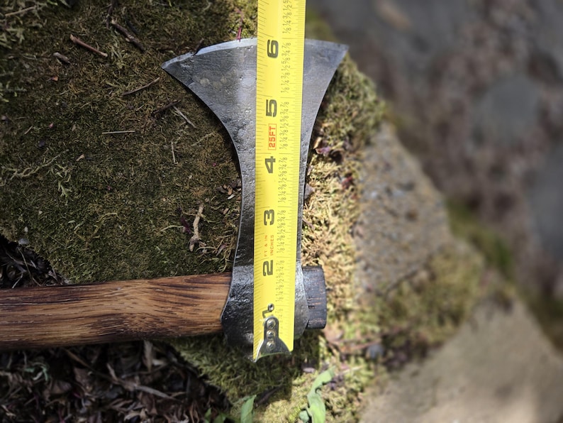Op de afbeelding: Een handbijl met een donkere metalen kop en een houten steel. De bijlkop is ongeveer 15 cm lang, gemeten met een gele rolmaat. De bijl ligt op een bed van groen mos.
