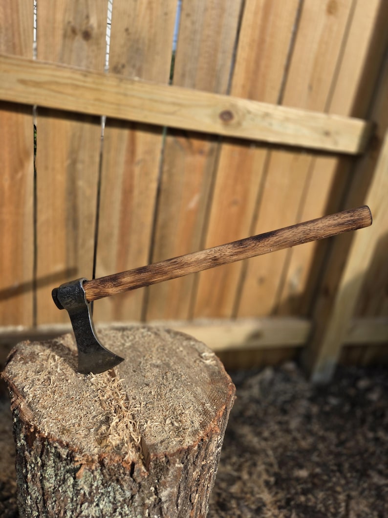 Op de afbeelding: Een handgemaakte bijl met een donkerhouten handvat en een zilveren blad, rustend op een boomstronk. De bijl is ontworpen om hout te hakken. De steel is ongeveer 46 cm lang.