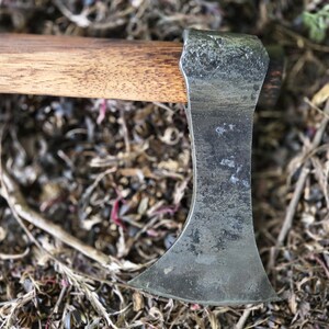 Op de afbeelding: Close-up van een kleine bijl met een houten steel en een verweerd, grijs metalen blad. De bijlkop rust tegen een achtergrond van droge bladeren en twijgen, wat het rustieke ontwerp benadrukt.