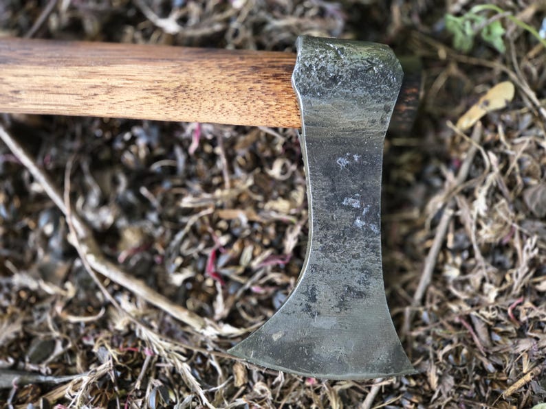 Op de afbeelding: Close-up van een kleine bijl met een houten steel en een verweerd, grijs metalen blad. De bijlkop rust tegen een achtergrond van droge bladeren en twijgen, wat het rustieke ontwerp benadrukt.