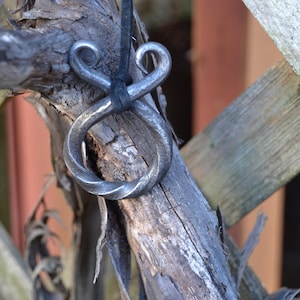 May include: A twisted metal pendant with a loop on the top, hanging from a black cord. The pendant is made of dark metal and has a rustic, handmade look.