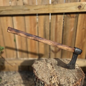 Op de afbeelding: Een handgemaakte bijl met een donkerhouten handvat en een gesmede stalen kop. De bijl rust op een boomstronk en toont zijn rustieke ontwerp. De handgreep is ongeveer 46 cm lang.