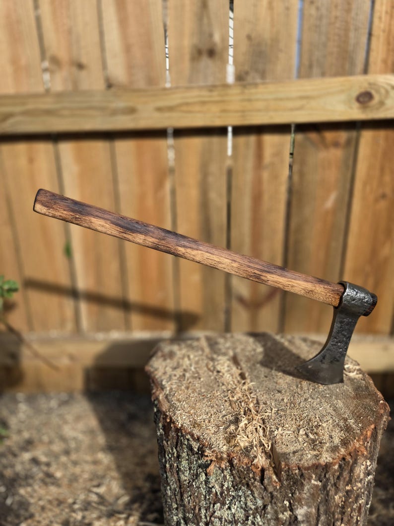 Op de afbeelding: Een handgemaakte bijl met een donkerhouten handvat en een gesmede stalen kop. De bijl rust op een boomstronk en toont zijn rustieke ontwerp. De handgreep is ongeveer 46 cm lang.