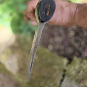 Op de afbeelding: Close-up van een tuingereedschap met een zwarte handgreep en een zilveren blad. Het gereedschap wordt in een hand gehouden, met een vage achtergrond van groen en steen.