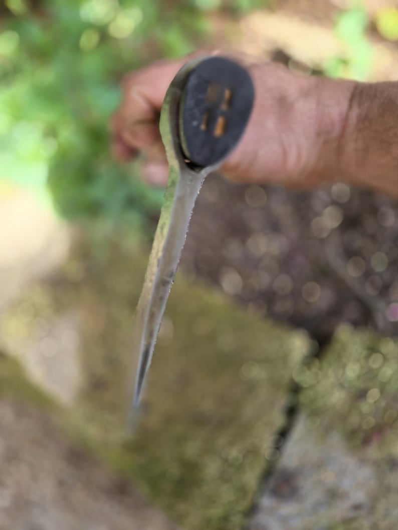 Op de afbeelding: Close-up van een tuingereedschap met een zwarte handgreep en een zilveren blad. Het gereedschap wordt in een hand gehouden, met een vage achtergrond van groen en steen.