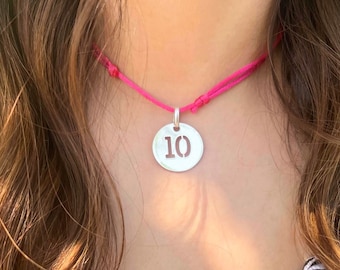 May include: A silver necklace with a round pendant featuring the number 10 cut out. The necklace is on a pink cord.