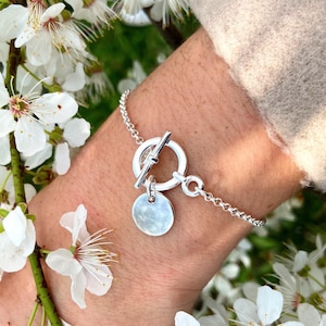 Puede incluir: Un brazalete de plata con cierre de palanca y un dije redondo. El brazalete está en la muñeca de una persona y el fondo es una flor blanca.