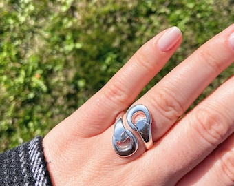 Puede incluir: Un anillo plateado con un diseño abierto único. El anillo presenta dos elementos curvos en forma de lágrima que se superponen, creando un aspecto abstracto y moderno. El anillo se lleva en un dedo, con un fondo verde y marrón.