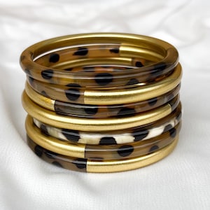 May include: Stack of six bangles. The bangles alternate between gold-colored and brown with black spots. One bangle has a white and black spotted pattern. The bangles are stacked on a white, textured surface.