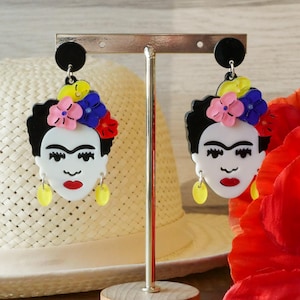 May include: A pair of Frida Kahlo-style earrings. Each earring depicts a white face with a red lip, black hair, and a floral headpiece with pink, blue, and yellow accents. The earrings have yellow teardrop-shaped dangles.