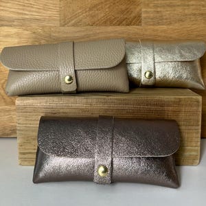 May include: Three leather eyeglass cases in taupe, gold, and bronze. Each case features a flap closure with a strap and a gold-coloured snap. The cases are displayed on a wooden surface.